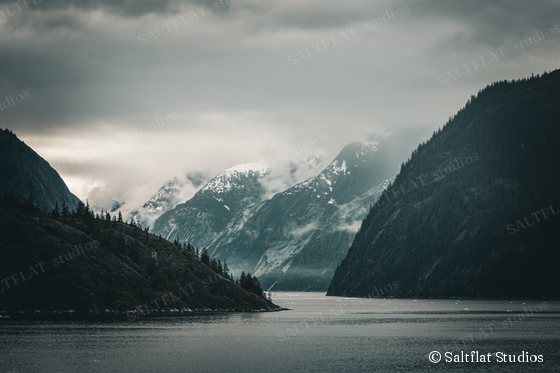 Dark ridges frame a calm fjord. Snowy peaks rise into low clouds beyond.
