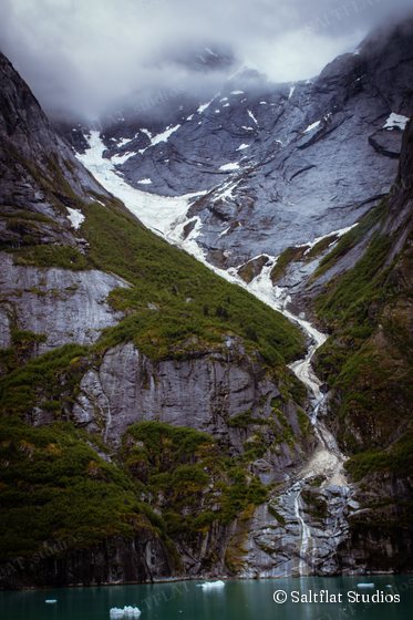 Meltwater runs from a glacier down steep gray cliffs into a green fjord dotted with small icebergs.