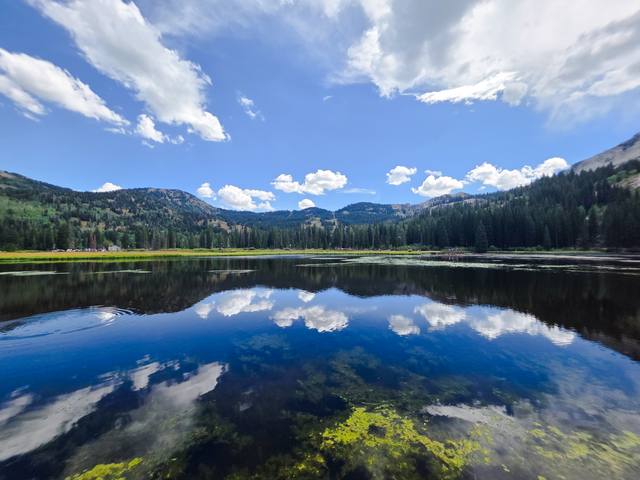 Clouds and green hills reflect in clear, still water.