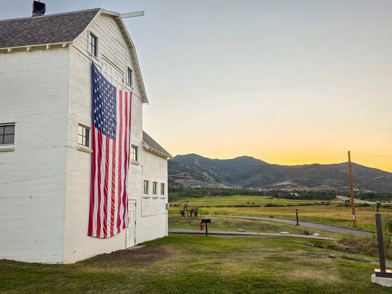 A white barn and flag below the mountains at sunset.