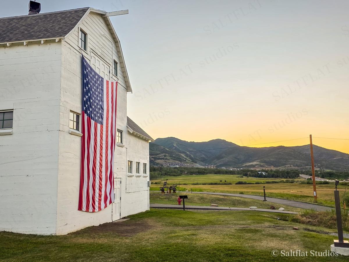 A white barn and flag below the mountains at sunset.